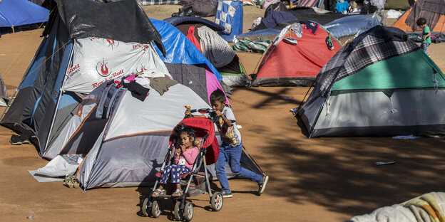 Ein Kind schiebt ein anderes auf einem Kinderwagen durch Zelte und Schlafmatten auf einem Gelände, das nahe der US-Grenze von Migranten als Schlafplatz benutzt wird.