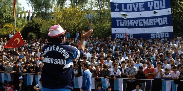 Ein Mann mit Türkiyemspor-Trikot steht auf einer Tartbahn vor einer Fankurve und klatscht in die Hände