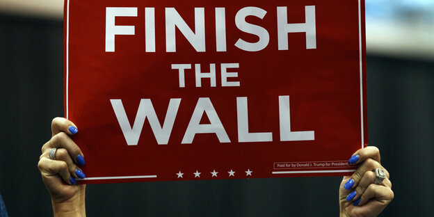 Eine Person hält ein Schild hoch, auf dem "Finish the Wall" steht Eine Person hält ein Schild hoch, auf dem "Finish the Wall" steht