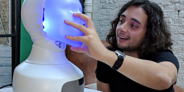 Ein Mann mit langen Haaren greift mit seiner Hand nach einem blau erleuchteten Kunstkopf