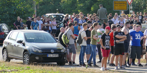 Eine große Gruppe junger Menschen steht auf einer Straße neben einem Auto und einem Ortsschild Eine große Gruppe junger Menschen steht auf einer Straße neben einem Auto und einem Ortsschild