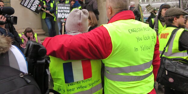 Erick Simon und Laurie Martin stehen Arm in Arm mit dem Rücken zur Kamera, auf ihren gelben Westen steht „English People: You look good in yellow"