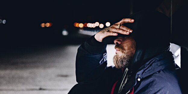 Ein Mann mit Vollbart und tätowierter Hand sitzt auf einer Straße. Ein Mann mit Vollbart und tätowierter Hand sitzt auf einer Straße.