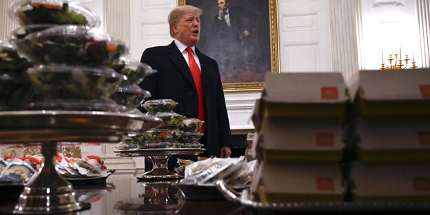 Trump steht hinter einem Tisch, der mit Burgern und Salaten in Plastikschalen vollgestellt ist Trump steht hinter einem Tisch, der mit Burgern und Salaten in Plastikschalen vollgestellt ist