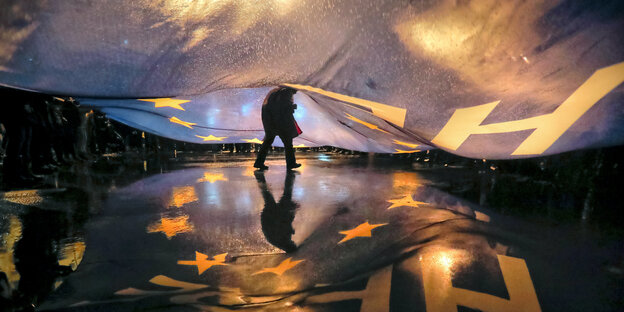 Eine Person läuft unter einem großen Banner durch. Auf dem nassen Boden spiegeln sich die Lichter Eine Person läuft unter einem großen Banner durch. Auf dem nassen Boden spiegeln sich die Lichter