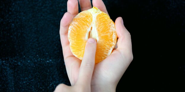 Eine halbe geschälte Orange in der einen Hand, zeigt der Zeigefinger der anderen Hand auf deren Mitte Eine halbe geschälte Orange in der einen Hand, zeigt der Zeigefinger der anderen Hand auf deren Mitte