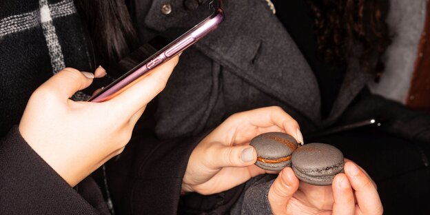 Mit einem rosa Smartphone werden zwei dunkelgrauen Macaron fotografiert Mit einem rosa Smartphone werden zwei dunkelgrauen Macaron fotografiert