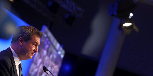 Markus Söder steht in einem dunklen Saal, fast versteckt in der unteren linken Bildhälfte Markus Söder steht in einem dunklen Saal, fast versteckt in der unteren linken Bildhälfte