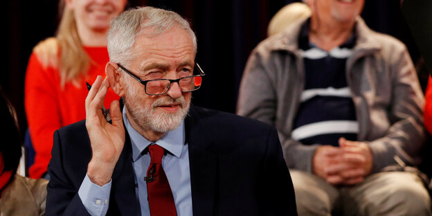 Jeremy Corbyn kneift die Augen zusammen und hält die Hand am Ohr. Hinter ihm sitzen Menschen Jeremy Corbyn kneift die Augen zusammen und hält die Hand am Ohr. Hinter ihm sitzen Menschen
