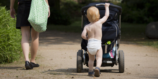 Ein Kleinkind, das nur eine Windel trägt, schiebt einen Kinderwagen in einem Park Ein Kleinkind, das nur eine Windel trägt, schiebt einen Kinderwagen in einem Park