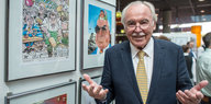 Dieter Hanitzsch steht vor eingerahmten Karikaturen und hebt seine Hände nach oben