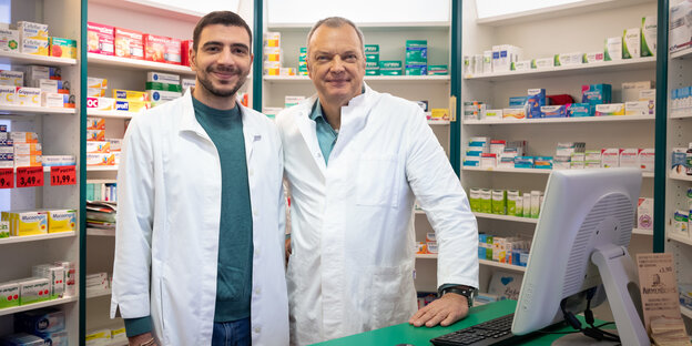 Zwei Apotheker stehen in Kitteln vor Regalen mit Medikamenten Zwei Apotheker stehen in Kitteln vor Regalen mit Medikamenten