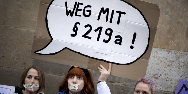 Junge Frauen, die ein Plakat mit der Aufschrft "Weg mit §219a" hochhalten Junge Frauen, die ein Plakat mit der Aufschrft "Weg mit §219a" hochhalten