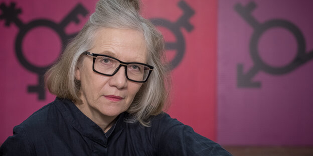 Porträtaufnahme von Cornelia Sollfrank vor lila-pinkem Hintergrund mit abstrakten Geschlechtersymbolen Porträtaufnahme von Cornelia Sollfrank vor lila-pinkem Hintergrund mit abstrakten Geschlechtersymbolen