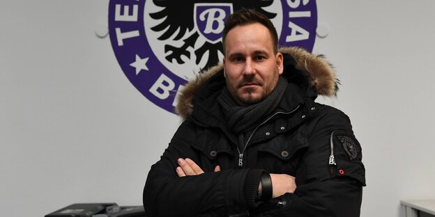 Jens Redlich steht mit verschränkten Armen vor dem Wappen von Tennis Borussia Berlin Jens Redlich steht mit verschränkten Armen vor dem Wappen von Tennis Borussia Berlin