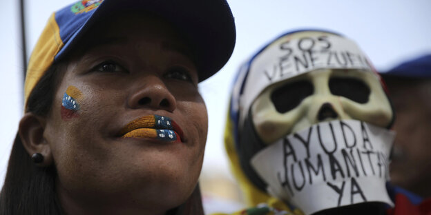 Eine Protestierende gegen Venezuelas Präsident Nicolás Maduroträgt ein Basecap und Lippenstift in den Farben der venezolanischen Flagge, neben ihr steht ein Demonstrant mit einer Totenkopfmaske Eine Protestierende gegen Venezuelas Präsident Nicolás Maduroträgt ein Basecap und Lippenstift in den Farben der venezolanischen Flagge, neben ihr steht ein Demonstrant mit einer Totenkopfmaske
