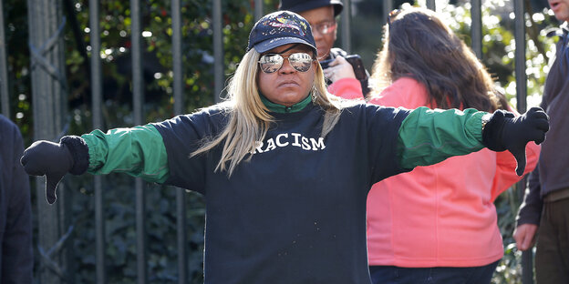 Eine schwarze Demonstrantin trägt ein Shirt mit der Aufschrift „Rassismus“ und zeigt die Daumen nach unten Eine schwarze Demonstrantin trägt ein Shirt mit der Aufschrift „Rassismus“ und zeigt die Daumen nach unten