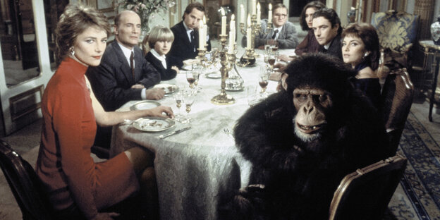 Bei dem Foto aus einem Film sitzen zahlreiche schick gekleidete Menschen am Essenstisch, gemeinsam mit einem Gorilla Bei dem Foto aus einem Film sitzen zahlreiche schick gekleidete Menschen am Essenstisch, gemeinsam mit einem Gorilla