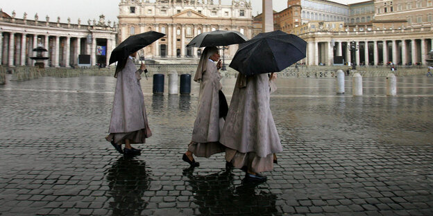 Drei Nonnen laufen bei Regen mit Regenschirmen über einen Platz Drei Nonnen laufen bei Regen mit Regenschirmen über einen Platz