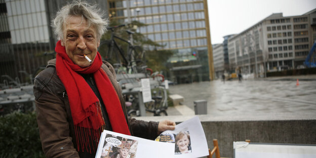 Artur Streit hat eine Zigarette im Mund und einen roten Schal um den Hals, in der Hand hält er zwei Bilder von Angela Merkel Artur Streit hat eine Zigarette im Mund und einen roten Schal um den Hals, in der Hand hält er zwei Bilder von Angela Merkel