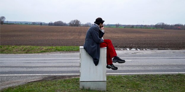 Ein Mann mit roter Hose und Hut sitzt auf einem Stromkasten. Im Hintergrund eine Straße und Landschaft