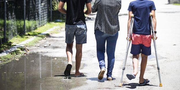 Drei Männer von hinten, die auf einem Weg laufen. Man sieht ihre Schultern und Köpfe nciht. Einer geht auf Krücken Drei Männer von hinten, die auf einem Weg laufen. Man sieht ihre Schultern und Köpfe nciht. Einer geht auf Krücken