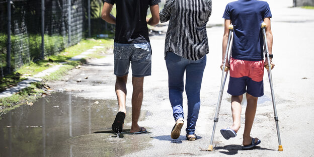 Drei Männer von hinten, die auf einem Weg laufen. Man sieht ihre Schultern und Köpfe nciht. Einer geht auf Krücken