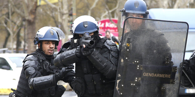 Drei Polizisten, einer von ihnen zielt mit einem Tränengasgewehr in Richtung Fotograf Drei Polizisten, einer von ihnen zielt mit einem Tränengasgewehr in Richtung Fotograf