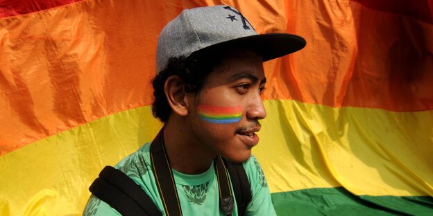 Ein Mann mit einer gemalten Regenbogenflagge auf der Backe steht vor einer Regenbogenflagge Ein Mann mit einer gemalten Regenbogenflagge auf der Backe steht vor einer Regenbogenflagge