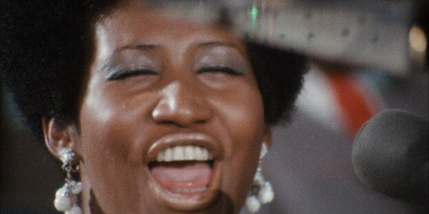 Das Gesicht der Sängerin Aretha Franklin. Sie trägt große Ohrringe, hat die Augen geschlossen und den Mund beim Singen weit geöffnet