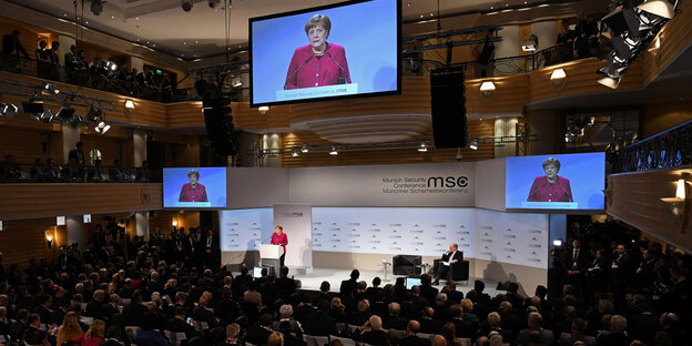 Der Saal der Sicherheitskonferenz mit Angela Merkel auf der Bühne auf drei großen Bildschirmen Der Saal der Sicherheitskonferenz mit Angela Merkel auf der Bühne auf drei großen Bildschirmen