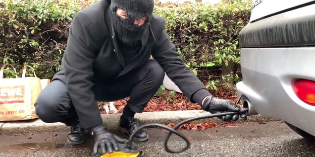 Ein Mann mit Sturmhaube zieht Abgase aus einem Auspuff in eine Luftpumpe