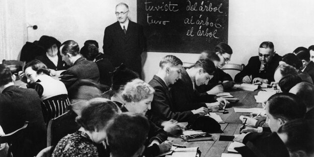 Schwarz-Weiß-Fotografie: An einem Tisch sitzen zahlreiche Personen und schreiben, an der Wand ist eine Tafel mit spanischen Wörtern angebracht Schwarz-Weiß-Fotografie: An einem Tisch sitzen zahlreiche Personen und schreiben, an der Wand ist eine Tafel mit spanischen Wörtern angebracht