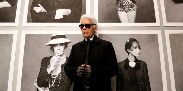 Karl Lagerfeld steht vor Porträtaufnahmen, er trägt eine schwarze Brille und schwarze Handschuhe Karl Lagerfeld steht vor Porträtaufnahmen, er trägt eine schwarze Brille und schwarze Handschuhe