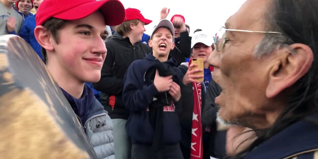 Ein Jugendlicher mit rotem Baseballcap steht einem indigenen Amerikaner gegenüber Ein Jugendlicher mit rotem Baseballcap steht einem indigenen Amerikaner gegenüber