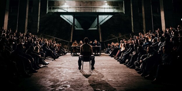 Filmszene: Ein Mann sitzt in einem Raum in der Mitte. Sehr viel leute sitzen um ihn herum und schauen ihn an Filmszene: Ein Mann sitzt in einem Raum in der Mitte. Sehr viel leute sitzen um ihn herum und schauen ihn an