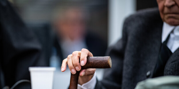 Greise Hand auf einem Stock, im Hintergrund sieht man verschwommen die dazugehörige Gestalt des angeklagten SS-Mannes Greise Hand auf einem Stock, im Hintergrund sieht man verschwommen die dazugehörige Gestalt des angeklagten SS-Mannes