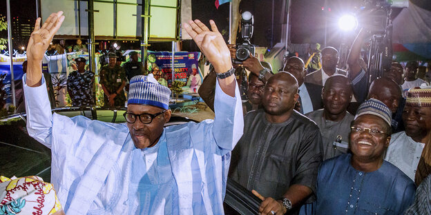 Buhari hebt jubelnd die Hände in die Höhe, im Hintergrund stehen mehrere Männer, darunter ein Fotograf und Polizisten. Buhari hebt jubelnd die Hände in die Höhe, im Hintergrund stehen mehrere Männer, darunter ein Fotograf und Polizisten.