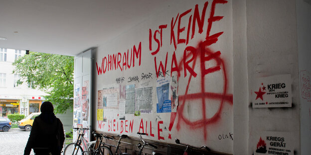 Frau geht an Graffiti gegen Immobilienspekulation an Häuserwand vorbei. Frau geht an Graffiti gegen Immobilienspekulation an Häuserwand vorbei.