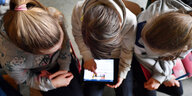 Drei Schulkinder, von oben fotografiert, sitzen nebeneinander. Das in der Mitte hält ein Tablet in der Hand.