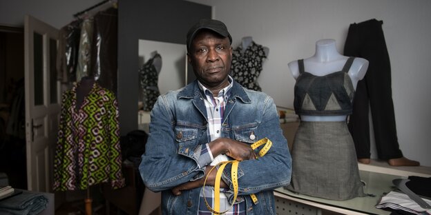 Ein dunkelhäutiger Mann in Jeansjacke mit Basecap auf dem Kopf und Metermaß in der Hand steht zischen Nähatelierpuppen, auf denen halb fertige Kleider hängen Ein dunkelhäutiger Mann in Jeansjacke mit Basecap auf dem Kopf und Metermaß in der Hand steht zischen Nähatelierpuppen, auf denen halb fertige Kleider hängen
