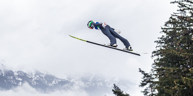 Ein Skispringer in der Luft. Im Hintergrund eine bewölkte Berglandschaft.