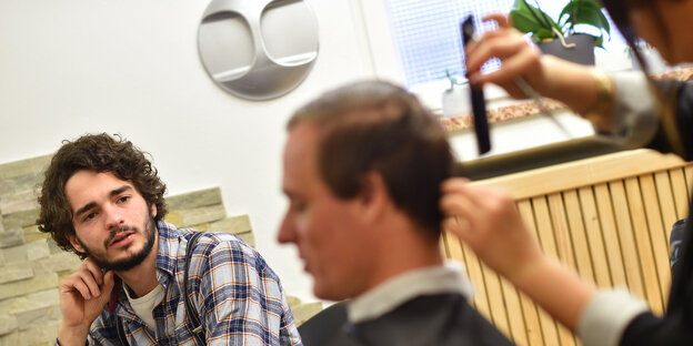 Einem Mann werden die Haare geschnitten, daneben sitzt ein anderer Mann Einem Mann werden die Haare geschnitten, daneben sitzt ein anderer Mann