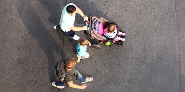 Zwei junge Männer gehen mit zwei Kindern über eine Straße. Eines der Kinder, ein kleines Mädchen, sitzt dabei in einem Kinderwagen. Zwei junge Männer gehen mit zwei Kindern über eine Straße. Eines der Kinder, ein kleines Mädchen, sitzt dabei in einem Kinderwagen.