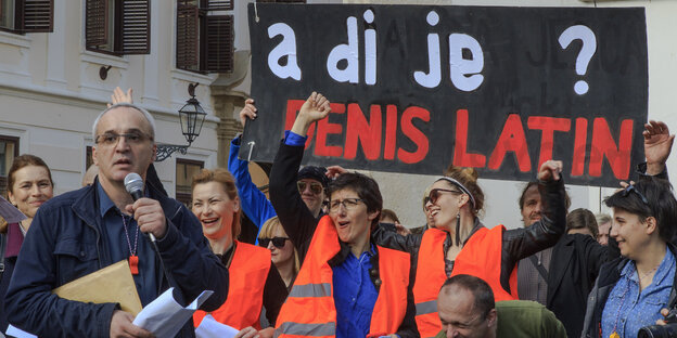 Ein Mann hat ein Mikrofon in der Hand, daneben DemonstrantInnen in orangefarbenen Warnwesten