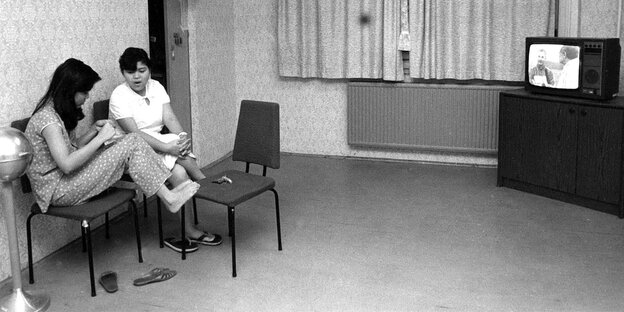 Zwei Frauen sitzen auf Stühlen in einem kargen Aufenthaltsraum, in einer Ecke läuft ein Fernseher Zwei Frauen sitzen auf Stühlen in einem kargen Aufenthaltsraum, in einer Ecke läuft ein Fernseher