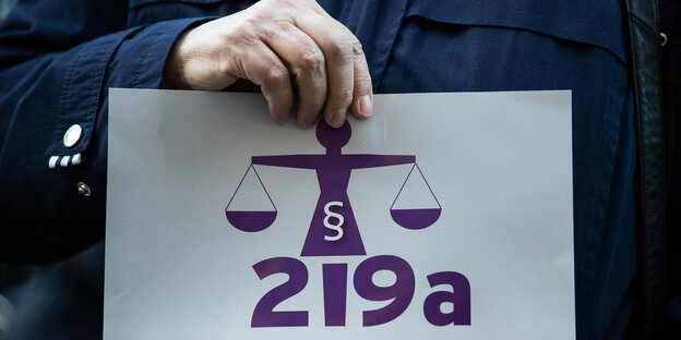 Eine Hand hält ein Schild auf dem in lila "§219a" und eine Waage zu sehen ist Eine Hand hält ein Schild auf dem in lila "§219a" und eine Waage zu sehen ist