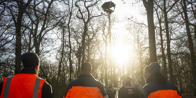 Menschen in orangefarbenen Warnwesten stehen im Hambacher Forst und schauen sich Baumhäuser an