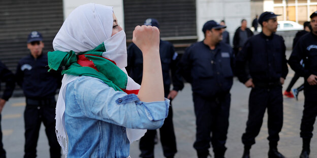 Eine Frau mit Kopftuch und algerischer Flagge um den Hals ballt die Hand zur Faust. Im Hintergrund stehen uniformierte Männer in einer Reihe. Eine Frau mit Kopftuch und algerischer Flagge um den Hals ballt die Hand zur Faust. Im Hintergrund stehen uniformierte Männer in einer Reihe.