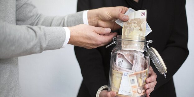 Eine Frau hält ein Glas mit Geld. EIn Mann steckt ihr mehr scheine rein Eine Frau hält ein Glas mit Geld. EIn Mann steckt ihr mehr scheine rein
